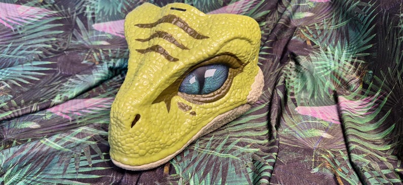 Puede incluir: Una hucha con forma de cabeza de dinosaurio amarilla con un ojo detallado y piel texturizada. El ojo es azul con una pupila oscura. La hucha est&aacute; sobre una tela con un estampado de hojas tropicales.