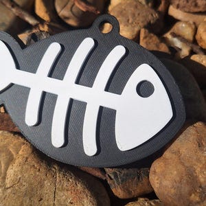 May include: A flat, black, fish-shaped tag featuring a white, raised skeletal fish bone design. The tag has a circular hole at the top for attachment, with a textured surface finish on both the black base and the white skeletal overlay.