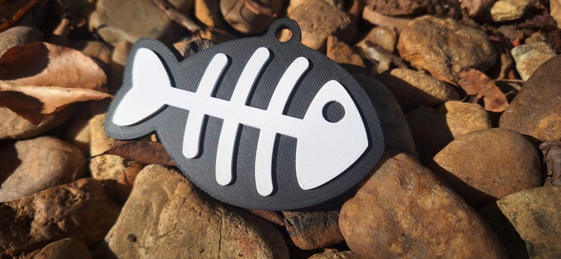 May include: A flat, black, fish-shaped tag featuring a white, raised skeletal fish bone design. The tag has a circular hole at the top for attachment, with a textured surface finish on both the black base and the white skeletal overlay.