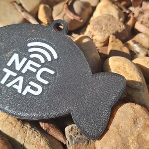 May include: A dark gray, fish-shaped NFC tag with a textured surface. The tag features a white graphic of a wireless signal icon above the white text "NFC TAP" printed on the center. A small circular hole is located at the top for attachment.
