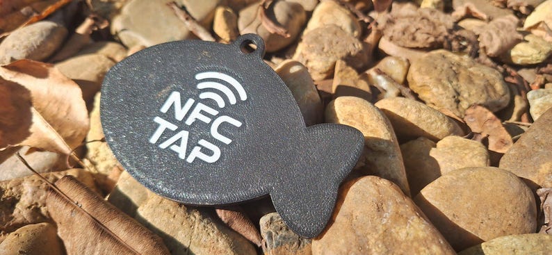 May include: A dark gray, fish-shaped NFC tag with a textured surface. The tag features a white graphic of a wireless signal icon above the white text "NFC TAP" printed on the center. A small circular hole is located at the top for attachment.