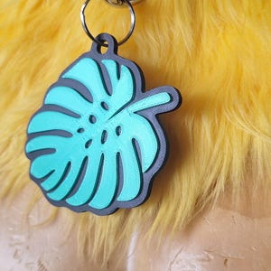 May include: A teal monstera leaf pendant with a black border, attached to a silver-colored metal ring. The pendant is positioned against a background of bright yellow faux fur, with a small circular sticker visible on the lower left side.