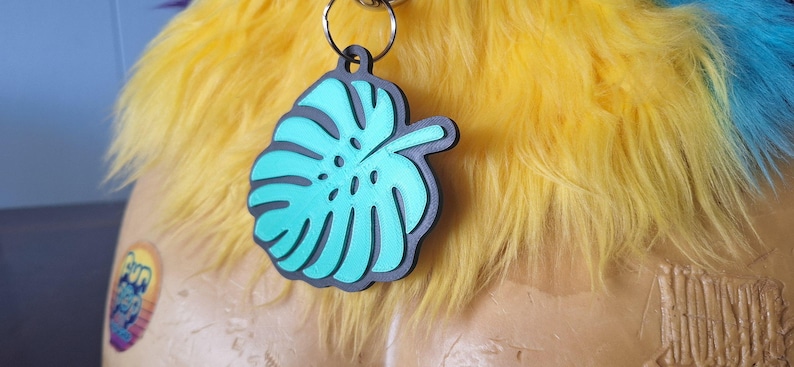 May include: A teal monstera leaf pendant with a black border, attached to a silver-colored metal ring. The pendant is positioned against a background of bright yellow faux fur, with a small circular sticker visible on the lower left side.
