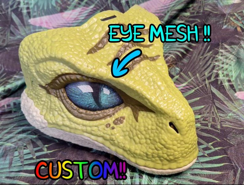Puede incluir: Una m&aacute;scara de dinosaurio personalizada en tonos amarillos y marrones, con un ojo azul detallado y malla ocular. La m&aacute;scara tiene una superficie texturizada y est&aacute; sobre un fondo con estampado de hojas tropicales. La palabra "CUSTOM!!" est&aacute; escrita en colores arco&iacute;ris.