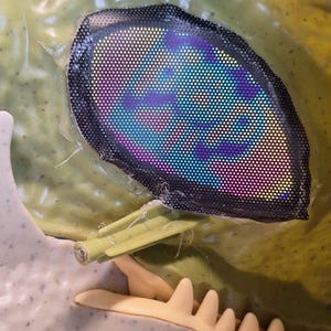 Puede incluir: Primer plano de una cabeza de dinosaurio de pl&aacute;stico verde con un ojo reflectante multicolor. El ojo est&aacute; enmarcado por una malla negra. La mand&iacute;bula y los dientes del dinosaurio son visibles.