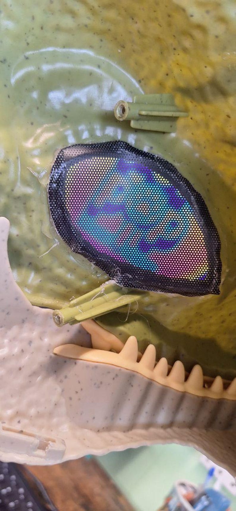 Puede incluir: Primer plano de una cabeza de dinosaurio de pl&aacute;stico verde con un ojo reflectante multicolor. El ojo est&aacute; enmarcado por una malla negra. La mand&iacute;bula y los dientes del dinosaurio son visibles.