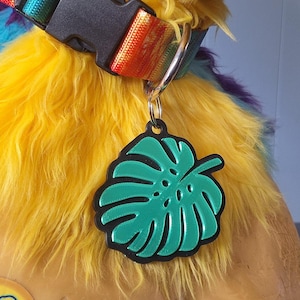 May include: A pet collar featuring a rainbow-colored nylon strap with a black plastic buckle and a metal D-ring. Attached to the D-ring is a green monstera leaf-shaped tag with a black border, resting against a yellow and purple faux fur collar.