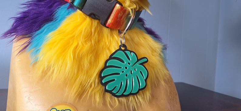 May include: A pet collar featuring a rainbow-colored nylon strap with a black plastic buckle and a metal D-ring. Attached to the D-ring is a green monstera leaf-shaped tag with a black border, resting against a yellow and purple faux fur collar.