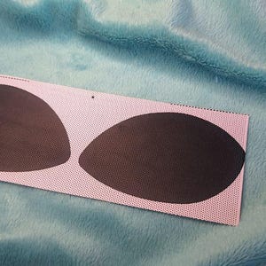 Puede incluir: Una tela rectangular rosa con dos almohadillas ovaladas de color marr&oacute;n oscuro. La tela tiene un patr&oacute;n de puntos y est&aacute; colocada sobre un fondo azul texturizado. Las almohadillas son lisas y opacas.