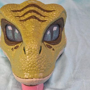 Puede incluir: Primer plano de un juguete con forma de cabeza de dinosaurio. El juguete es principalmente verde-amarillo con rayas marrones en la cabeza. Tiene ojos grandes y azules y una lengua rosa. El juguete probablemente sea de pl&aacute;stico.