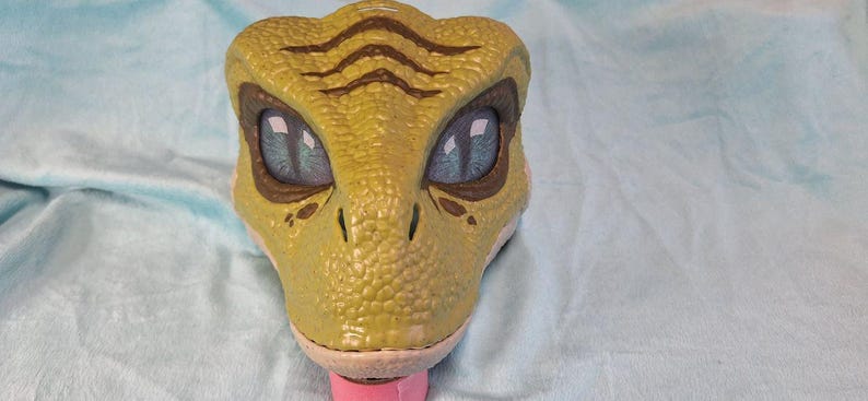 Puede incluir: Primer plano de un juguete con forma de cabeza de dinosaurio. El juguete es principalmente verde-amarillo con rayas marrones en la cabeza. Tiene ojos grandes y azules y una lengua rosa. El juguete probablemente sea de pl&aacute;stico.