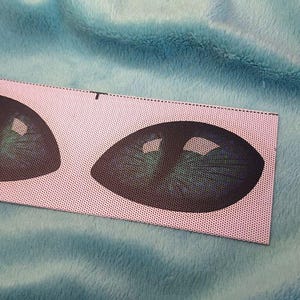 Puede incluir: Un parche rectangular de tela con dos ojos de gato estilizados, con detalles verdes y negros. El parche est&aacute; sobre un fondo azul claro y suave.
