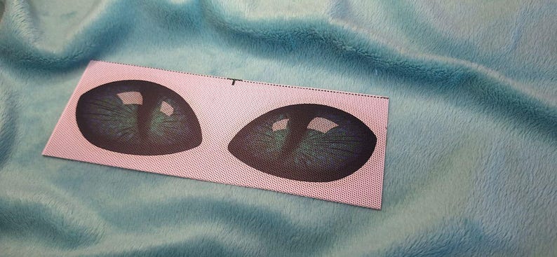 Puede incluir: Un parche rectangular de tela con dos ojos de gato estilizados, con detalles verdes y negros. El parche est&aacute; sobre un fondo azul claro y suave.