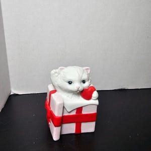 May include: A white ceramic kitten figurine holding a red heart, emerging from a pink gift box tied with a red ribbon. The kitten has blue eyes and is a Valentine's Day or love-themed decorative item.