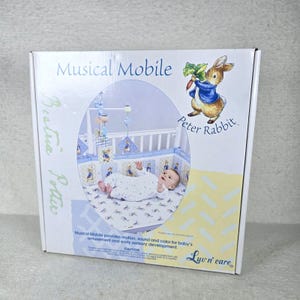 May include: A white rectangular product box for a Peter Rabbit musical mobile. The box features the text Musical Mobile, Beatrix Potter, and Luv n care. An illustration of Peter Rabbit and a photo of a baby in a crib are displayed.