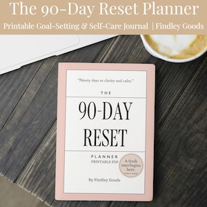 Pode incluir: Um planejador de redefinição de 90 dias rosa e branco com o texto "The 90-Day Reset Planner" e "Printable Goal-Setting & Self-Care Journal". O planejador está em uma superfície de madeira escura ao lado de um laptop e uma xícara de café.