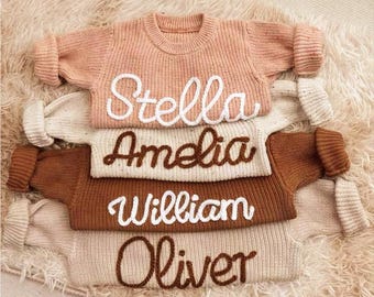May include: Four knitted sweaters stacked on top of each other. The sweaters are in different colors, including beige, brown, and pink. Each sweater has a name embroidered on it in white thread: Stella, Amelia, William, and Oliver.