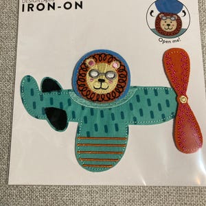 May include: An interactive iron-on patch featuring a lion wearing goggles inside a teal airplane with a brown propeller. The packaging includes the text Design Loft Iron-On and Open me! with a circular graphic showing the lion head lifting up.