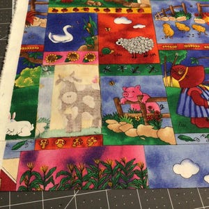 May include: A colorful fabric panel featuring farm animal illustrations. The design includes a swan, sheep, cow, pig, and a character in a red hat and blue dress. The fabric has a patchwork style with a variety of scenes and a white rabbit.