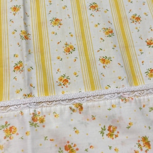 Könnte beinhalten: Weißer Stoff mit gelben vertikalen Streifen und floralen Mustern. Der Stoff weist ein sich wiederholendes Muster aus gelben und orangenen Blumen und kleinen grünen Blättern auf. Am unteren Rand ist eine weiße Spitzenborte zu sehen.
