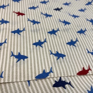 May include: White fabric with vertical gray stripes and a repeating pattern of blue and red shark silhouettes. The sharks are evenly spaced across the fabric, creating a playful and nautical design.