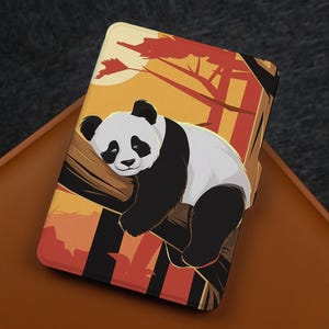 May include: A rectangular e-reader case featuring a graphic illustration of a black and white panda resting on a tree branch. The background displays a stylized sunset with warm orange, yellow, and deep red tones, depicting a forest setting.