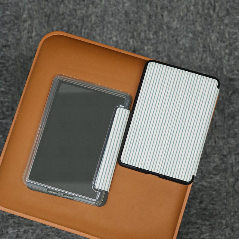 Puede incluir: Una funda protectora transparente y una funda a rayas para un lector electr&oacute;nico se muestran sobre una superficie marr&oacute;n. La funda presenta un patr&oacute;n de finas l&iacute;neas azules verticales sobre un fondo blanco. La pantalla del lector es oscura.