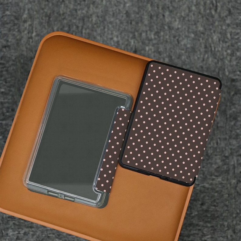 May include: A clear tablet case and a brown polka dot tablet cover are displayed on a brown surface. The tablet cover has a pattern of small, light pink dots. The tablet is visible inside the clear case.