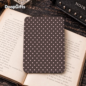 May include: A rectangular e-reader cover featuring a dark brown background with a repeating pattern of small, light pink polka dots. The cover is positioned flat on top of an open book with visible printed text on the pages, showing chapter fourteen.