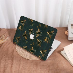 Op de afbeelding: Een laptop met een donkergroene hoes met een patroon van goud en zwarte bijen en bladeren. De laptop is open en toont het Apple-logo. De hoes ligt op een ronde, geweven placemat.