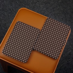 May include: Two brown cases with white polka dots are displayed on a brown surface. The cases are rectangular and appear to be for a tablet or e-reader. The background is a dark gray.