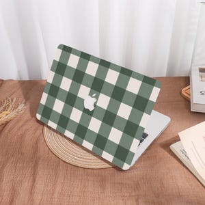 May include: A laptop with a green and white checkered pattern case. The case features the Apple logo in white. The laptop is open, revealing the keyboard. The case is resting on a woven placemat on a brown surface.