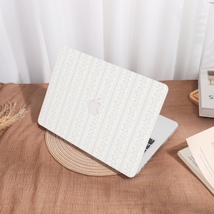 May include: A laptop with a white cover featuring a floral pattern with vertical stripes in light blue. The laptop is resting on a woven placemat on a brown surface. The Apple logo is visible on the laptop.
