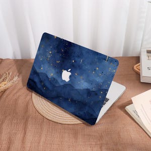Op de afbeelding: Een laptop met een marineblauwe hoes met een aquarelontwerp van een sterrenhemel met gouden accenten. Het Apple-logo is prominent in het wit weergegeven. De laptop staat open op een bruin oppervlak.