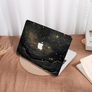 Op de afbeelding: Een zwarte laptophoes met een sterrenhemelontwerp. De hoes heeft gouden sterren en een wit Apple-logo. Het ontwerp heeft een aquareleffect met gouden accenten. De laptop ligt op een geweven mat.