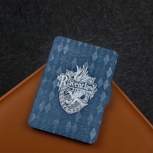 Könnte beinhalten: Eine blaue Hülle mit dem Ravenclaw-Wappen aus der Harry-Potter-Reihe. Das weiße Wappen zeigt einen Adler, ein Banner mit "Ravenclaw" und einen Schild. Die Hülle hat ein Rautenmuster und liegt auf einer braunen Oberfläche.