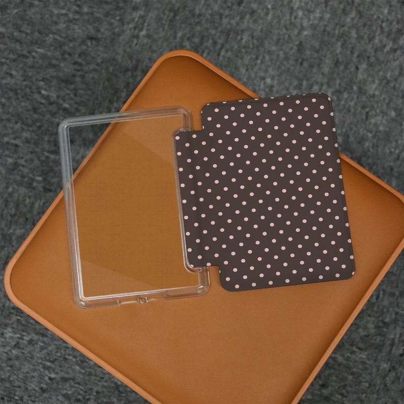 May include: A clear, rectangular phone case next to a brown phone case with a white polka dot pattern. The cases are on a brown surface.
