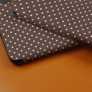 May include: Close-up of two brown cases with a repeating pattern of small, white polka dots. The cases have black edges and are slightly overlapping, creating a layered effect. The background is a solid, warm orange color.