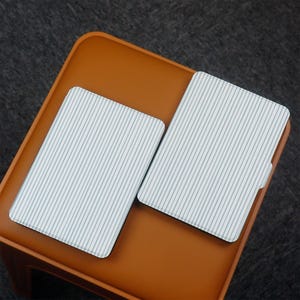 Puede incluir: Dos posavasos rectangulares con fondo blanco y finas rayas azules verticales. Los posavasos est&aacute;n sobre una superficie marr&oacute;n, posiblemente una mesita o taburete. El fondo es gris oscuro.