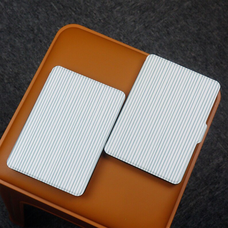Puede incluir: Dos posavasos rectangulares con fondo blanco y finas rayas azules verticales. Los posavasos est&aacute;n sobre una superficie marr&oacute;n, posiblemente una mesita o taburete. El fondo es gris oscuro.