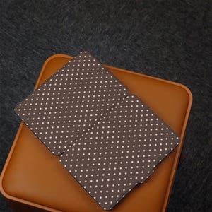 May include: A brown tablet case with a white polka dot pattern, open and resting on a brown leather-like surface. The case is rectangular with rounded corners and a smooth finish. The background is a dark gray, textured surface.