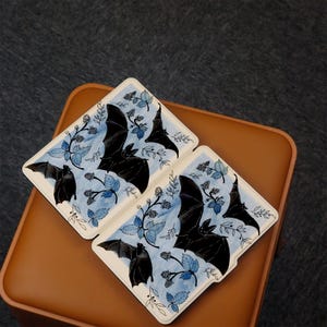 Puede incluir: Dos tarjetas blancas con ilustraciones azules y negras de murci&eacute;lagos volando sobre un patr&oacute;n de hojas azules y bayas negras. Las tarjetas est&aacute;n sobre una superficie marr&oacute;n.