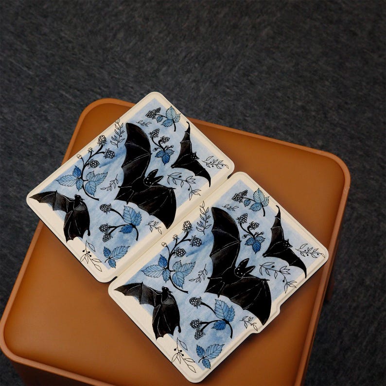 Puede incluir: Dos tarjetas blancas con ilustraciones azules y negras de murci&eacute;lagos volando sobre un patr&oacute;n de hojas azules y bayas negras. Las tarjetas est&aacute;n sobre una superficie marr&oacute;n.