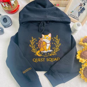 May include: Black hooded sweatshirt with a yellow embroidered design of a cat wearing a backpack and a laurel wreath. The text "QUEST SQUAD" is embroidered below the cat. The left sleeve has the word "BROCCOLI" embroidered on it.