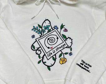 Könnte beinhalten: Ein weißer Kapuzenpullover mit einer zentralen Stickerei, die einen quadratischen Rahmen mit einer Spirale zeigt, umgeben von Ranken, Blättern und Blumen. Der Text "life is strange" steht unter der Spirale. Auf dem Ärmel steht "This action will have consequences..."
