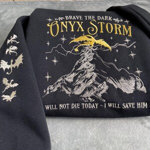 Op de afbeelding: Zwart sweatshirt met gouden borduurwerk met een draak, een berg en sterren. De tekst "BRAVE THE DARK ONYX STORM" en "WILL NOT DIE TODAY - I WILL SAVE HIM" is geborduurd op het sweatshirt.