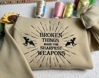 May include: Tan colored sweatshirt with black embroidered text that reads "Broken Things Make The Sharpest Weapons" with two black dragon silhouettes on either side of the text.