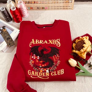 Könnte beinhalten: Roter Pullover mit einem schwarzen Drachen und einem Blumendesign. Der Text "ABRAXOS GARDEN CLUB" ist in Gold auf der Vorderseite gestickt.
