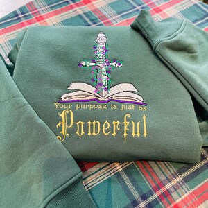 May include: A green sweatshirt with an embroidered design of a book with a sword through it. The text "Your purpose is just as Powerful" is embroidered below the book.