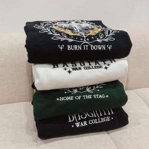 May include: A stack of four black and white sweatshirts with embroidered designs. The top sweatshirt has a black background with a white design of a hand holding a burning torch with the text "BURN IT DOWN". The second sweatshirt has a white background with the text "BASGIATH WAR COLLEGE" in black. The third sweatshirt has a green background with the text "HOME OF THE STAG" in white. The bottom sweatshirt has a black background with the text "BASGIATH WAR COLLEGE" in white.
